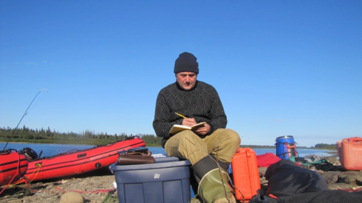 A man wearing a tuque, heavy sweater, waterproof pants and rubber boots is seating ona rock making notes in a book with a lake in the background.