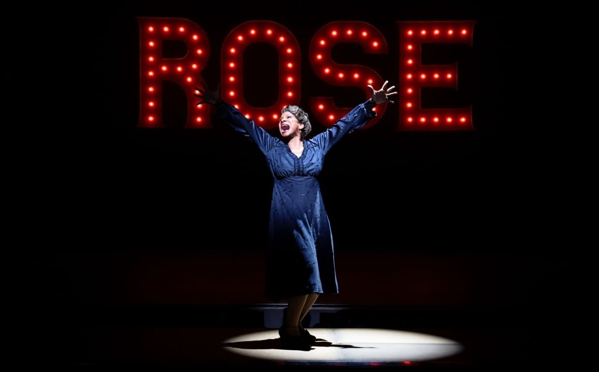 audra A woman stands in a spotlight with her arms spread wide under red lights spelling out the name "ROSE".