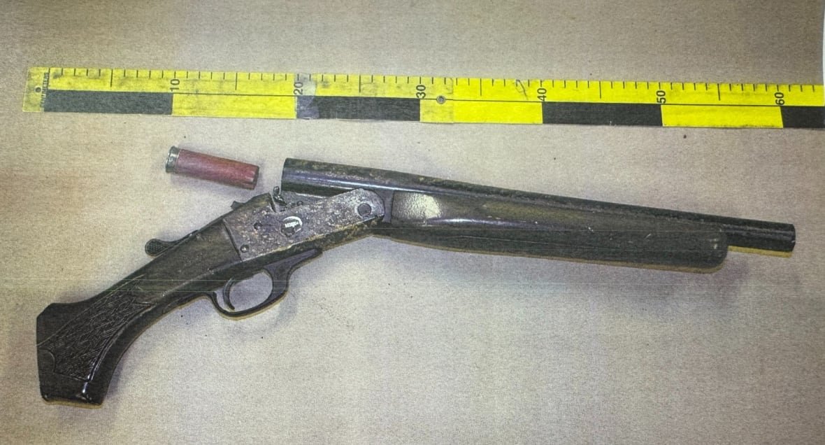 A firearm and a shell casing on a table with a measuring tape beside them.