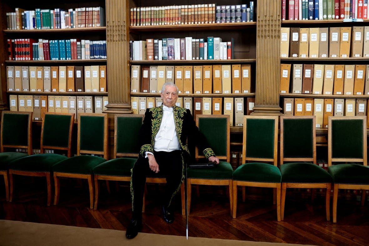people-mario-vargas-llosa A man in a green velvet embroidered suit sits in a high-back wooden chair with a green velvet seat and back in a row of identical chairs, in front of several bookcases.