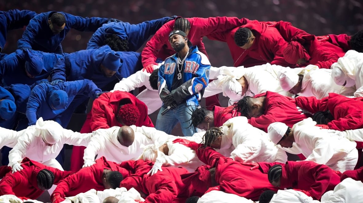 super-bowl-football-photo-gallery A man stands with his arms folded over his stomach in a crowd of dancers who are all bent over and holding one another by the back. The dancers are wearing either red, white or blue outfits, and are arranged so that the man who is standing appears to be standing inside of the American flag.