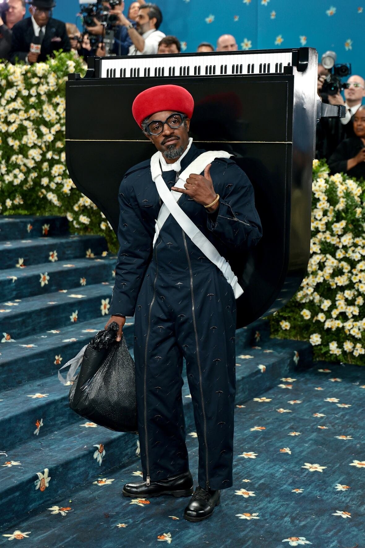 andre-3000-2025-met-gala A man wearing a blue jumpsuit and a miniature grand piano on his back poses for a photo.