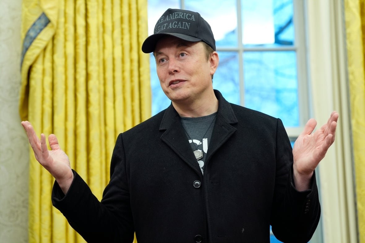 musk A clean-shaven man wearing a dark blazer, T-shirt and baseball cap extends his arms while speaking indoors.