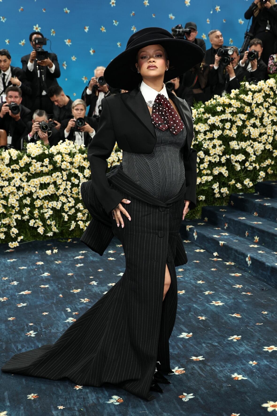 rihanna-2025-met-gala A woman in a black and grey corseted gown and a black hat poses for a photo.