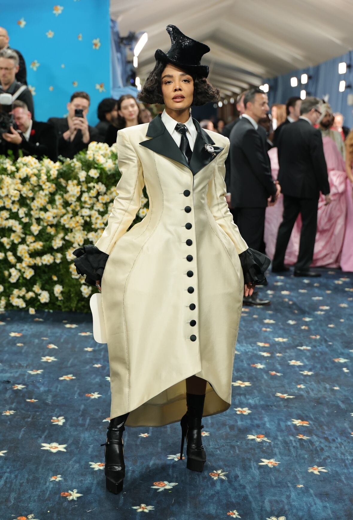 tessa-thompson-2025-met-gala A woman wearing a cream and black tailored coat dress and a small black top hat poses for a photo