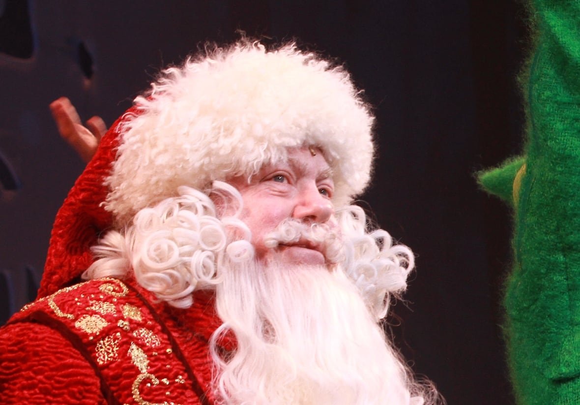 106880617 A man in a Santa suit and two men in elf costumes wave.