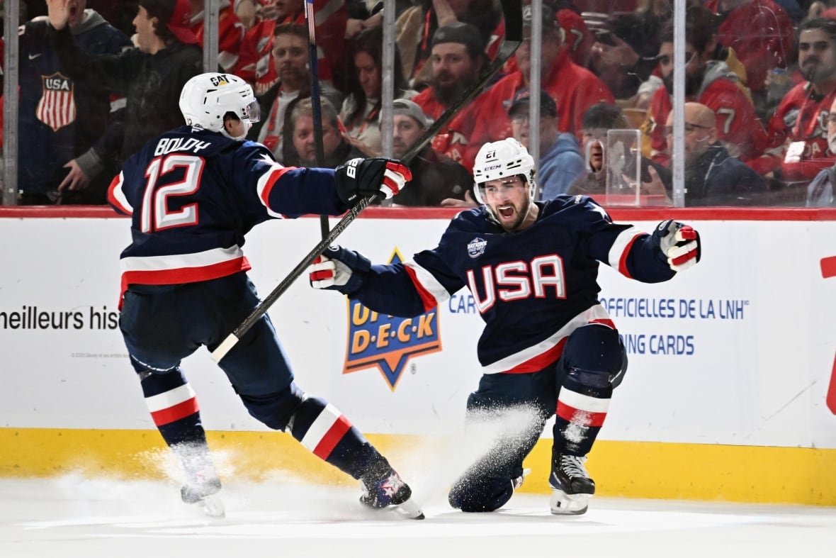 2199897256 Two male ice hockey players lift their arms in celebration on the ice as fans watch from behind the glass.