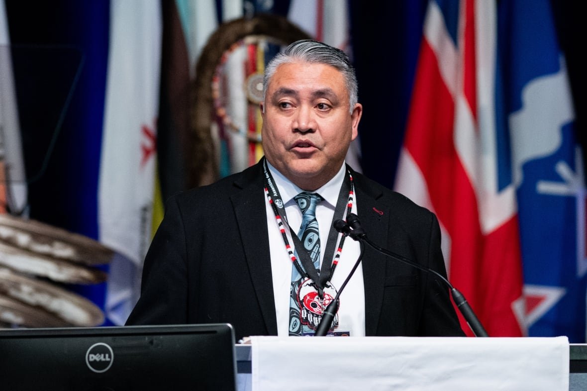 afn-assembly-20241204-1 A man speaks into a microphone with flags behind him.