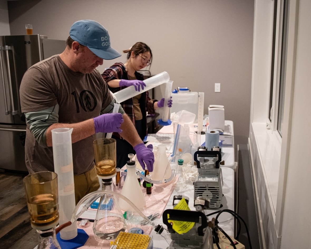 andrew-bramburger-algae-samples a man in a ball cap in the foreground and a woman with glasses in the background work with test tubes and other equipment, processing and analyzing samples of algae