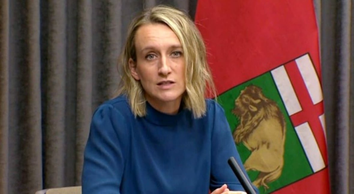 dr-joss-reimer A woman in a blue sweater speaks at a news conference while seated at a table, in front of a Manitoba flag.