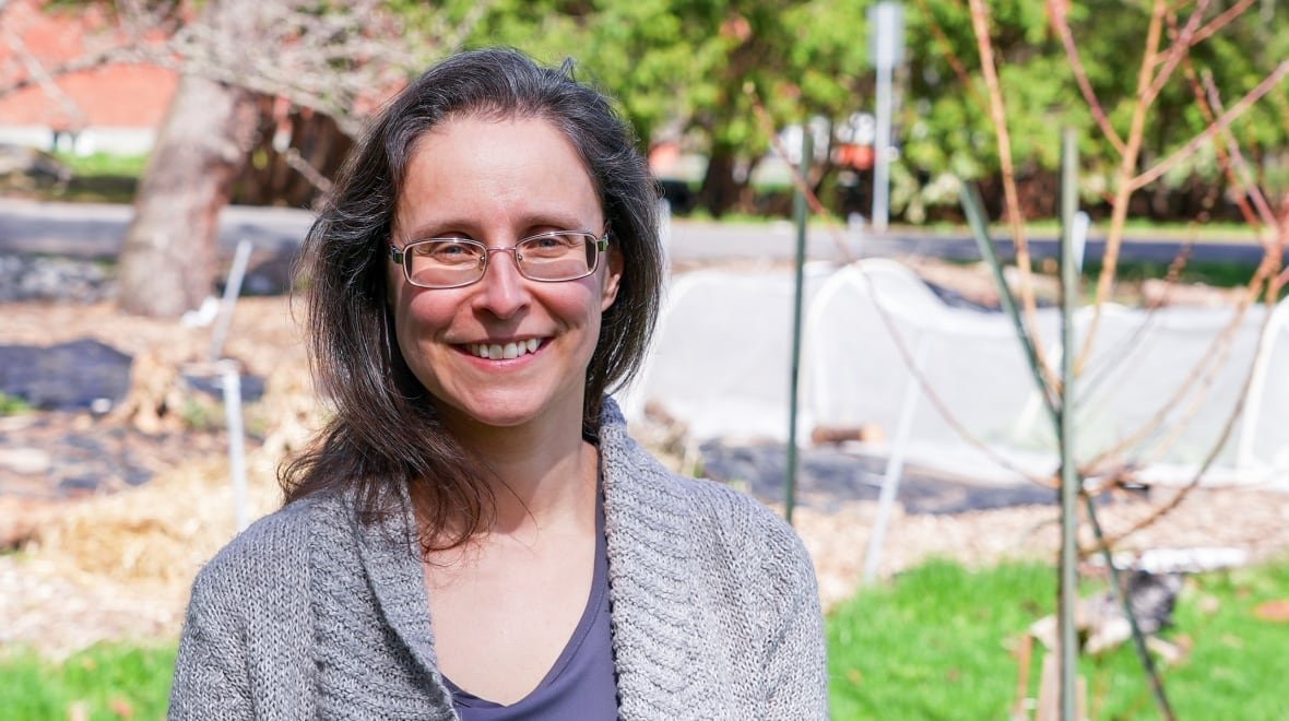 helene-hebert-gardening-consultant-aylmer-gatineau-quebec A woman with long hair and glasses smiles in front of a garden. A low, white tent-like tunnel structure is visible on the right-hand side of the image, behind a thin tree.