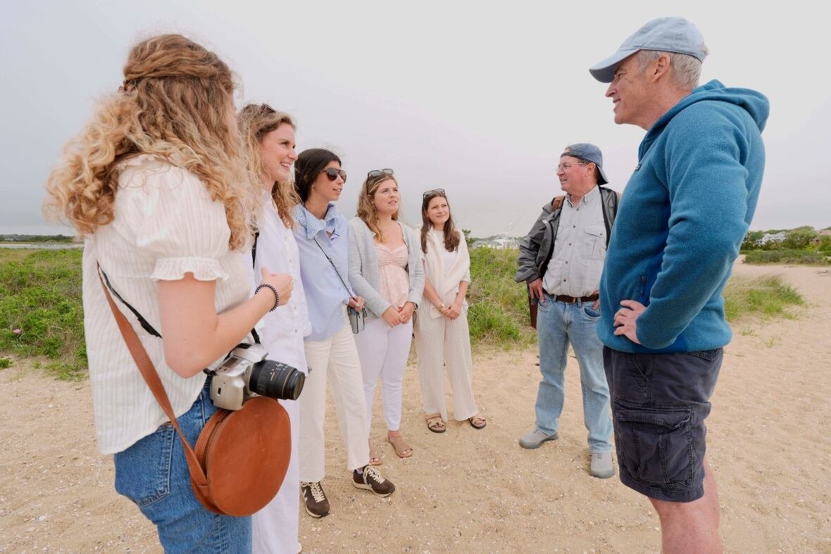jaws-anniversary-shark-swim A man in a hoodie, shorts and ball cap stands on a beach, his hands on his hips, as he speaks five women in summerwear, all lined up side-by-side.