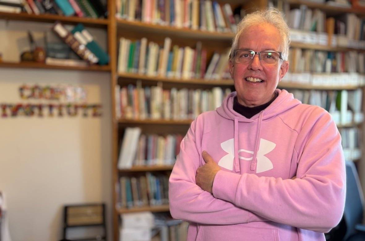 joe-byrne An older man with white hair and glasses in a pink hoodie stands in front of a large bookcase.