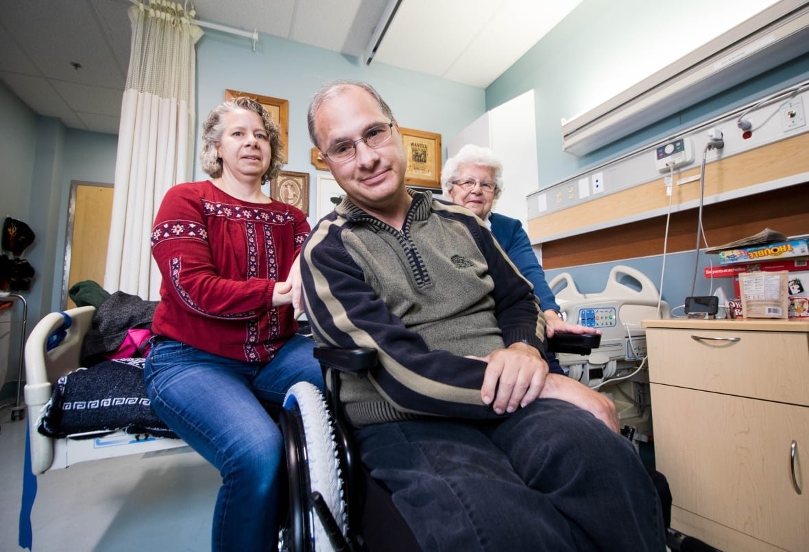ken-rekowski-hospital-renfrew-family-kareen-irene-brain-injury A man sits in a wheelchair with his arms crossed in a hospital room, flanked by two other women.