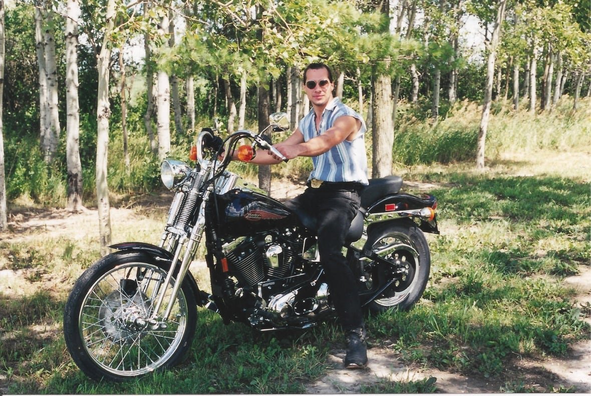 ken-rekowski-motorcycle-harley A man in sunglasses and a sleeveless shirt sits on a motorcycle on a sunny summer day.