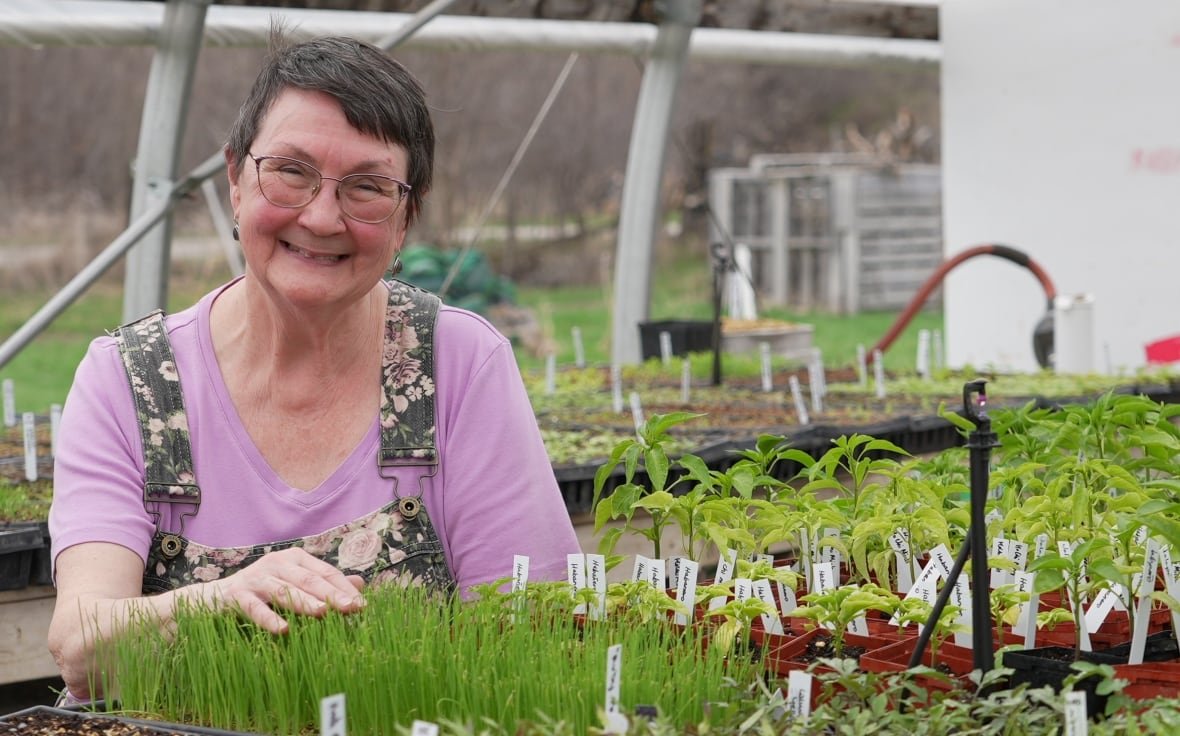 maureen-russell-growing-together-community-farm-2025-1 A woman with short hair smiles as she brushes her palm over leek seedlings. She wears a pink tshirt and floral-patterned overalls.