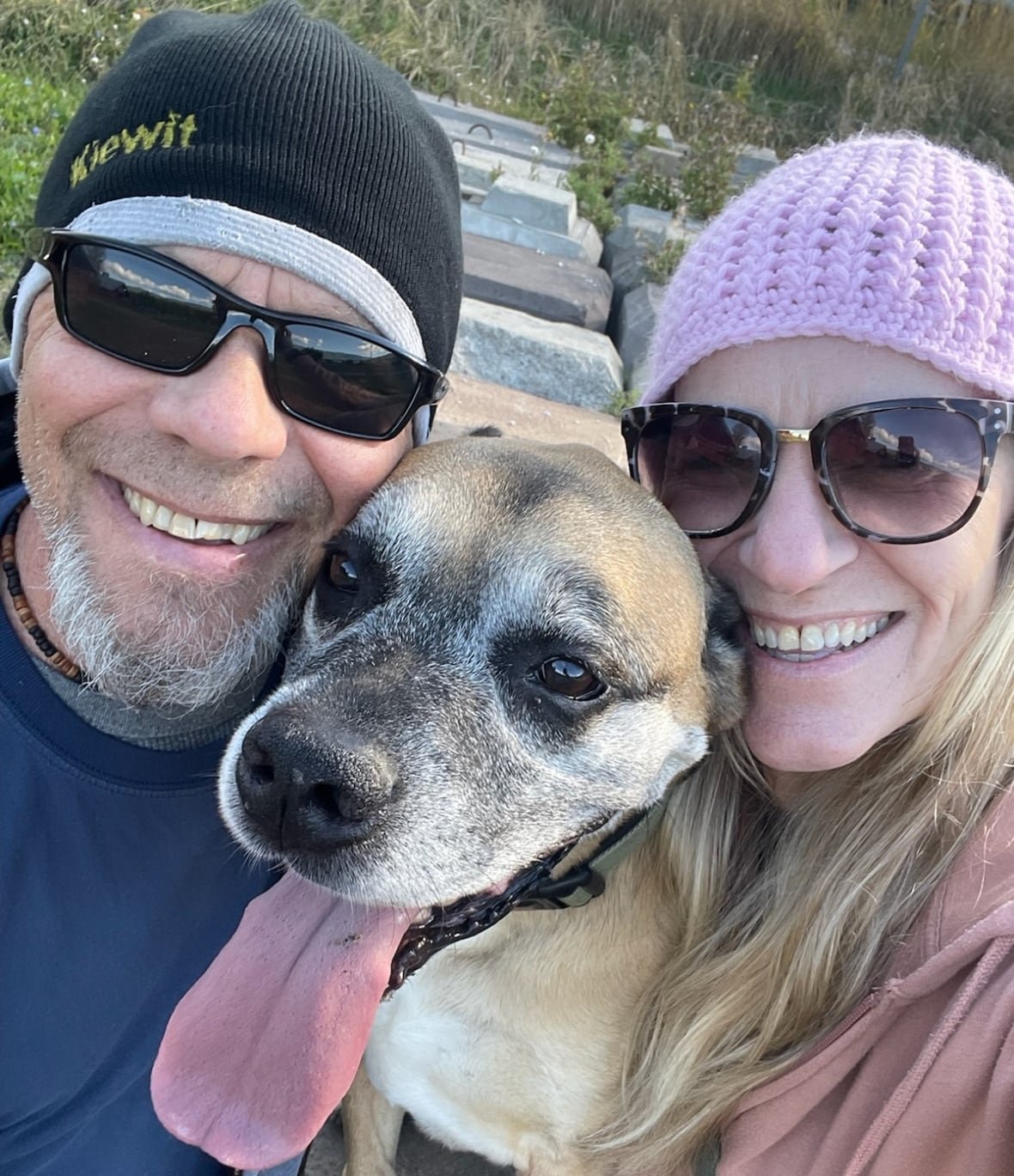 richard-anderson-first-person-with-michelle-harvey A smiling man and woman, both wearing knitted hats, pose with a dog in a selfie photograph.