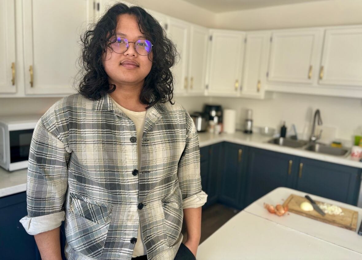 saugat-shrestha Man with glasses and long curly brown hair wears a grey and white plaid shirt as he stands in a kitchen.