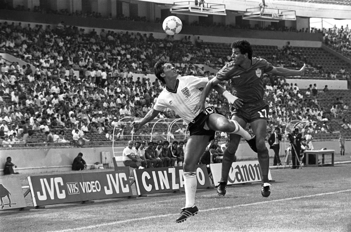 soccer-bbc-lineker Two male soccer players jump to head the ball on a pitch in this black and white photo from 1986. Behind them is the goal net and fans are seen in the stands.