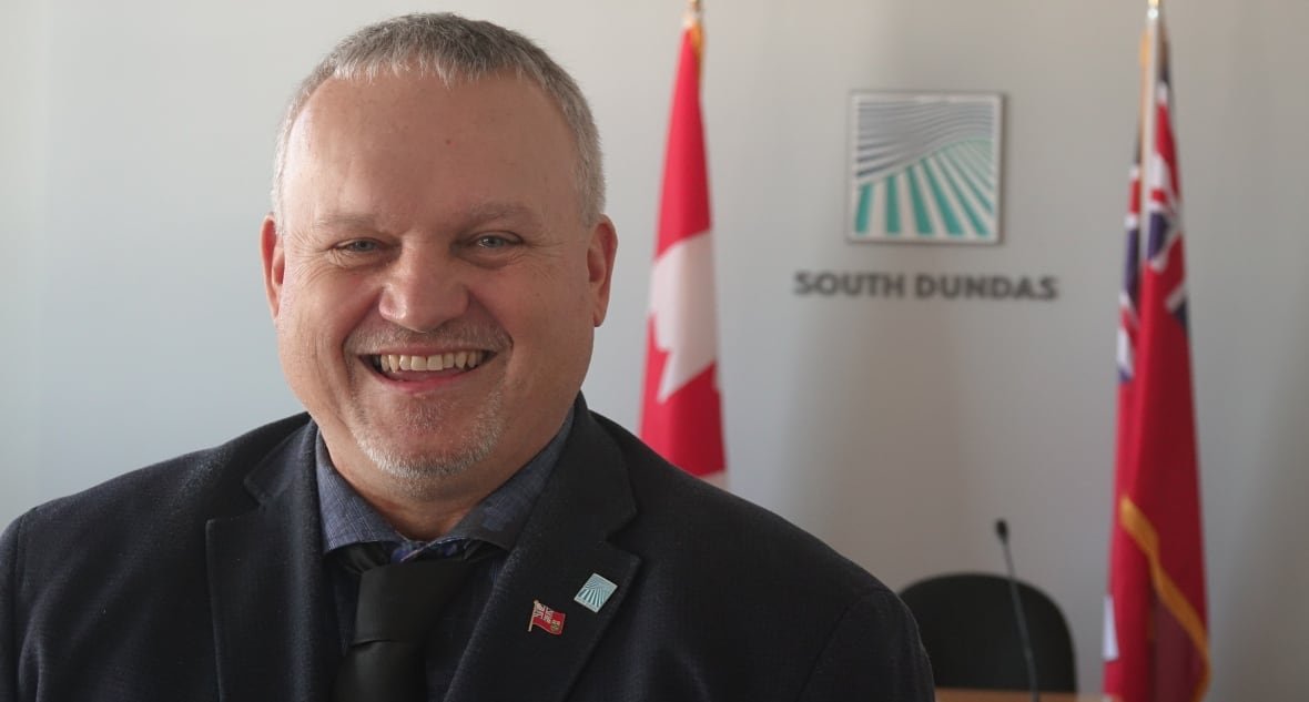 south-dundas-mayor-jason-broad A man with short grey hair, wearing a jacket and tie, stands in front of the Canadian and Ontario flags, along with a blue and green logo for the Municipality of South Dundas.