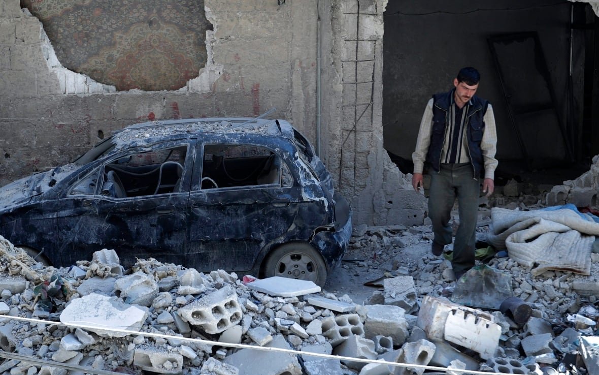 syria-survivors A man walks among the rubble of a destroyed building, with a car nearby.