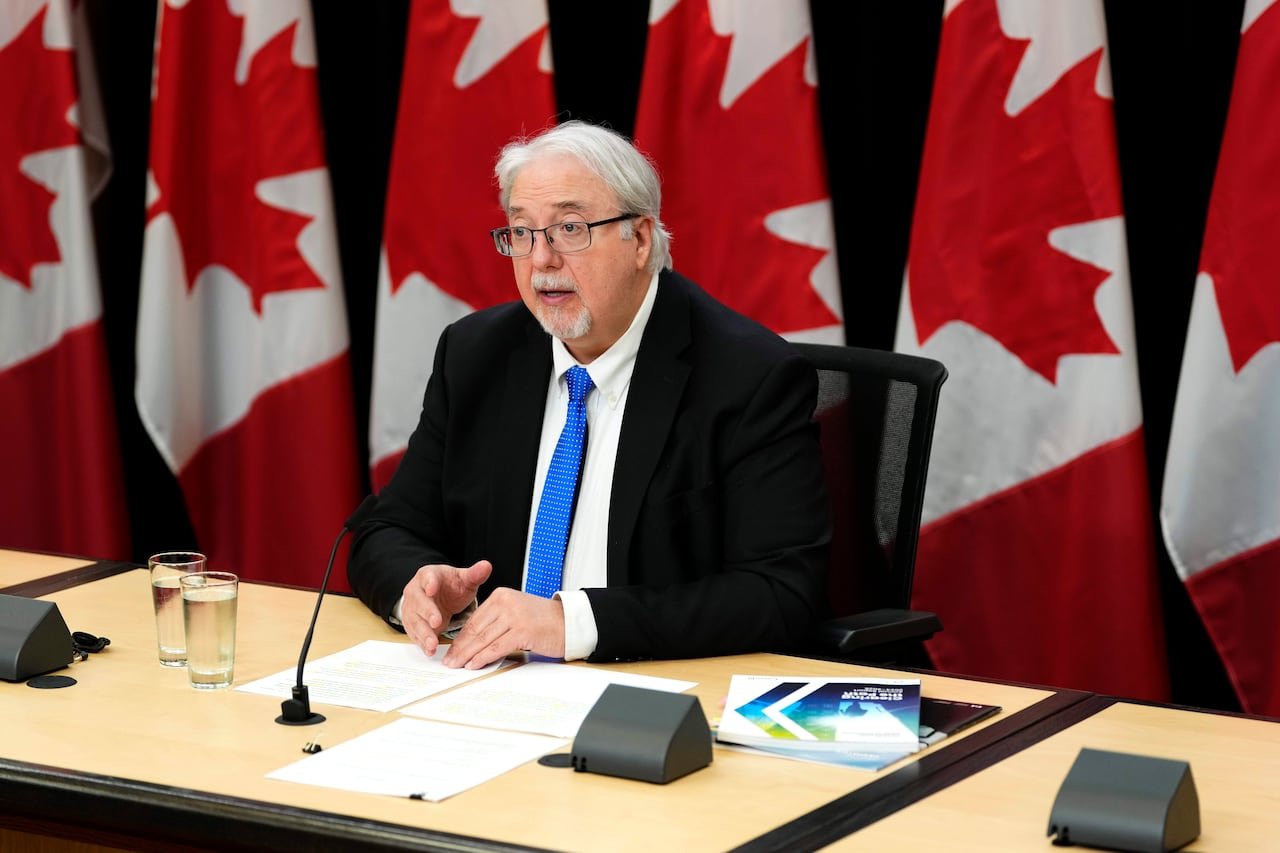 A man in a black suit and blue tie speaks into a microphone while holding some papers. A row of Canadian flags are draped in the background.
