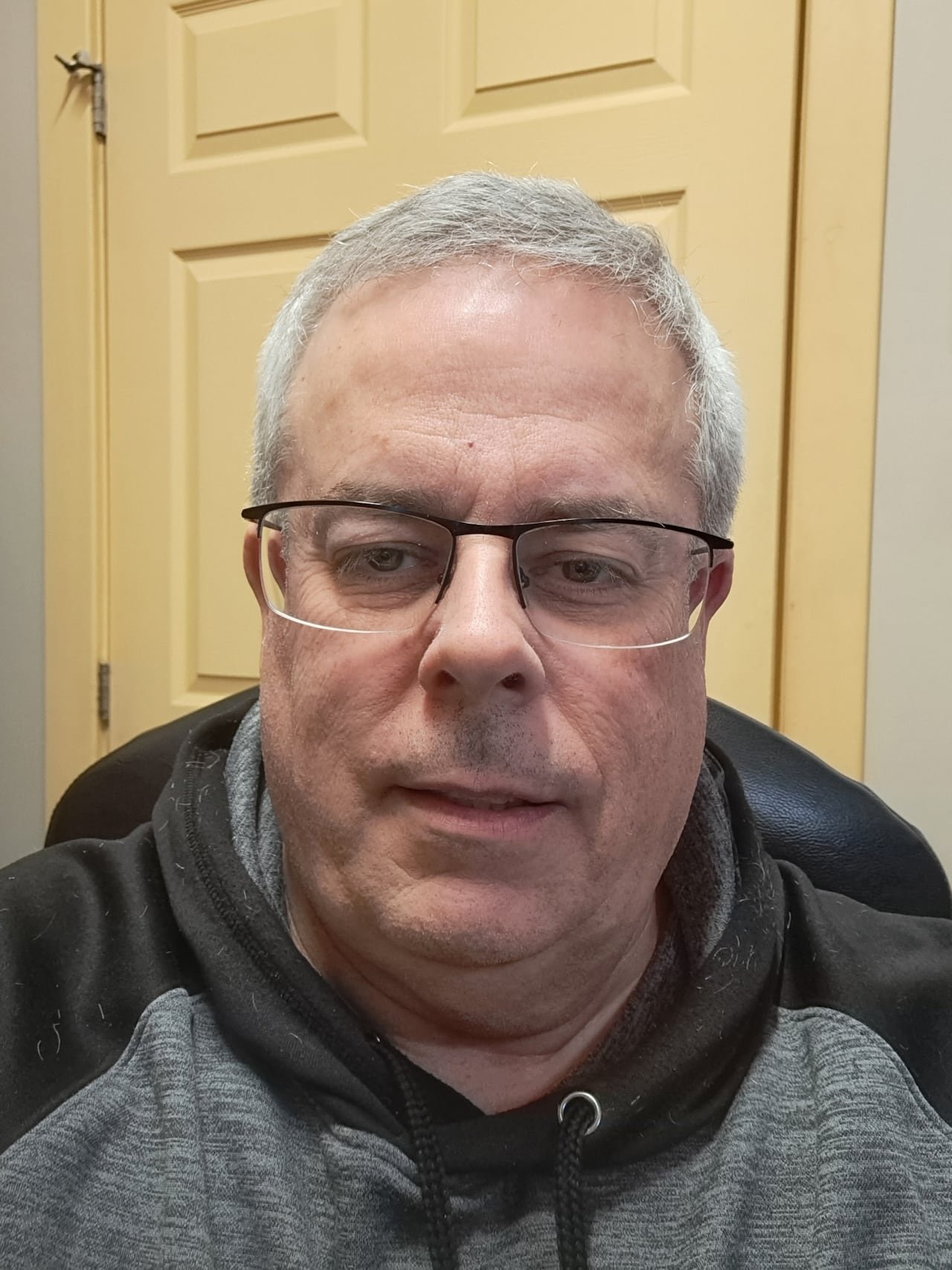 A white man with white hair, wears glasses. Wearing a black and grey hoodie.