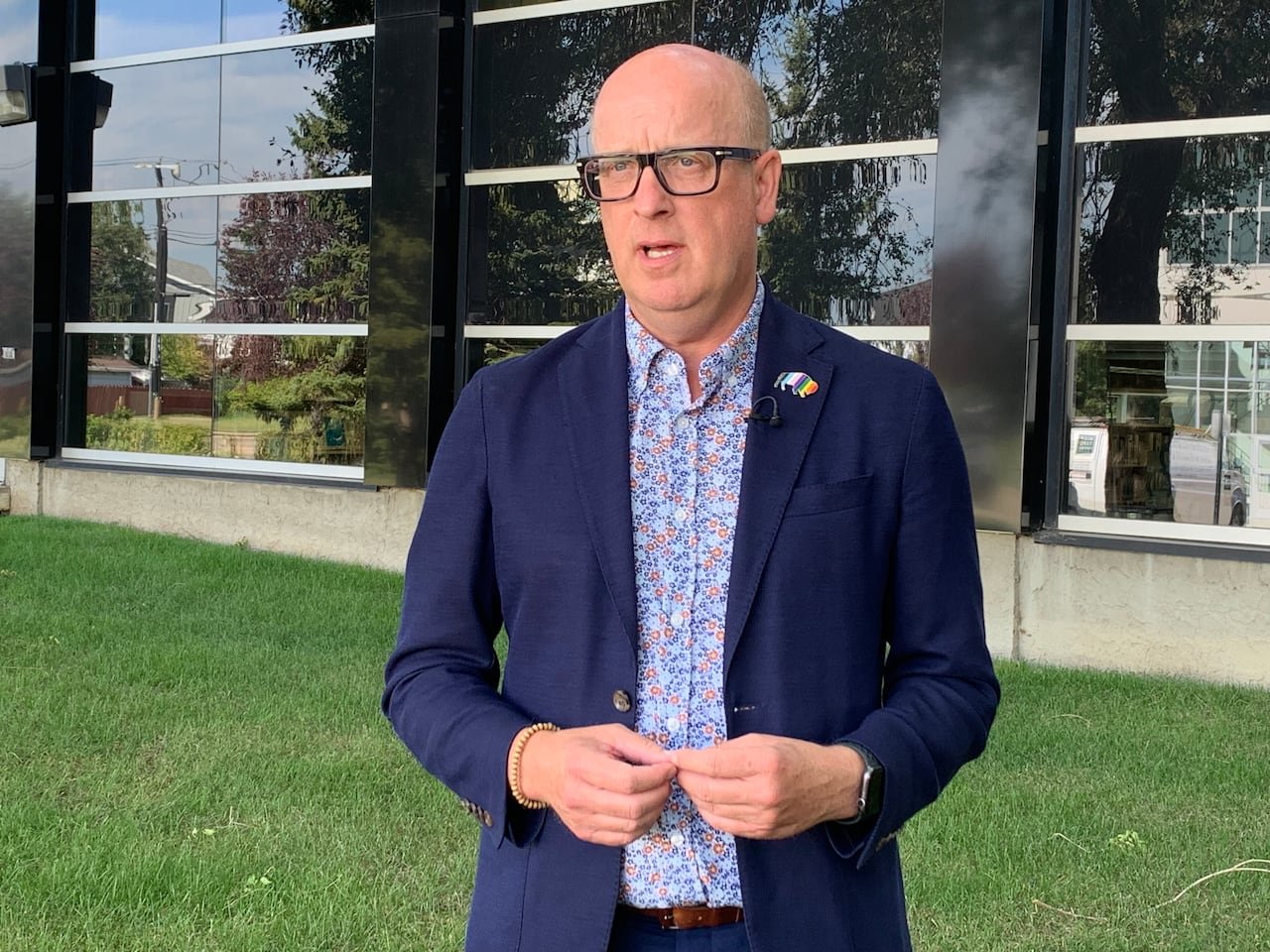 A man in a navy blazer and flowered dress shirt stands in front of a glass building on a green lawn.
