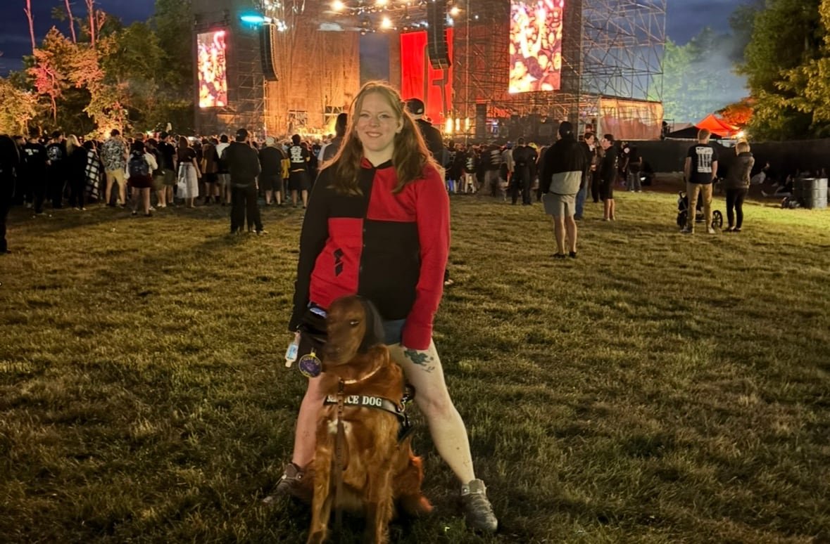 default-14 A woman stands in the crowd at an outdoor concert, her golden retriever standing between her legs.