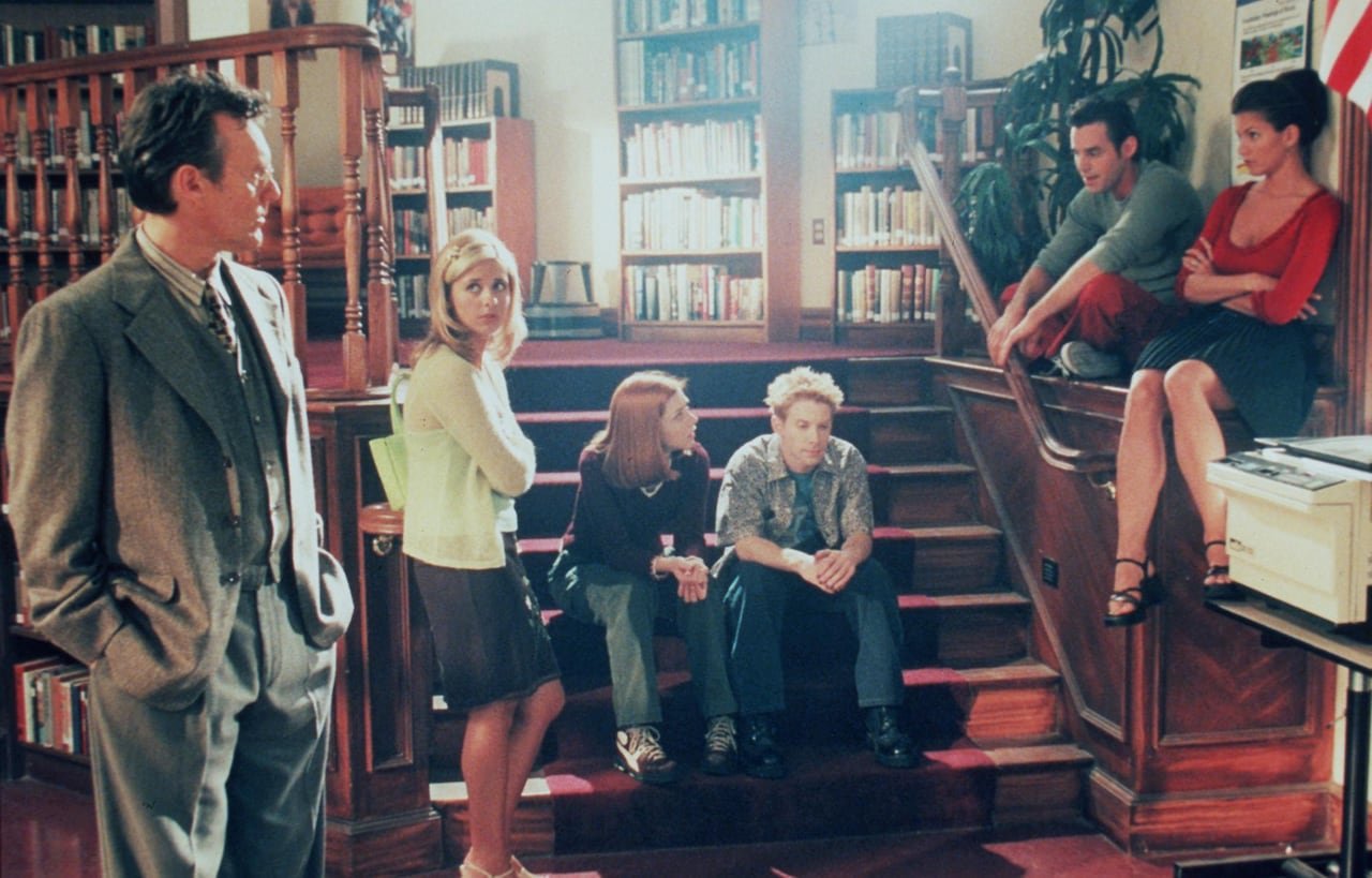 default-566 An adult man in a suit at left stands looking at a group of teens, standing and seated, in a library.