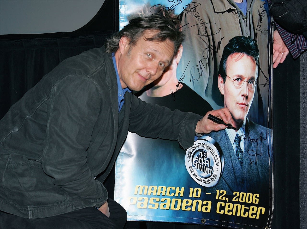 default-567 A smiling man looks over his shoulder as he pauses signing his autograph on a large poster that includes a photo of him.