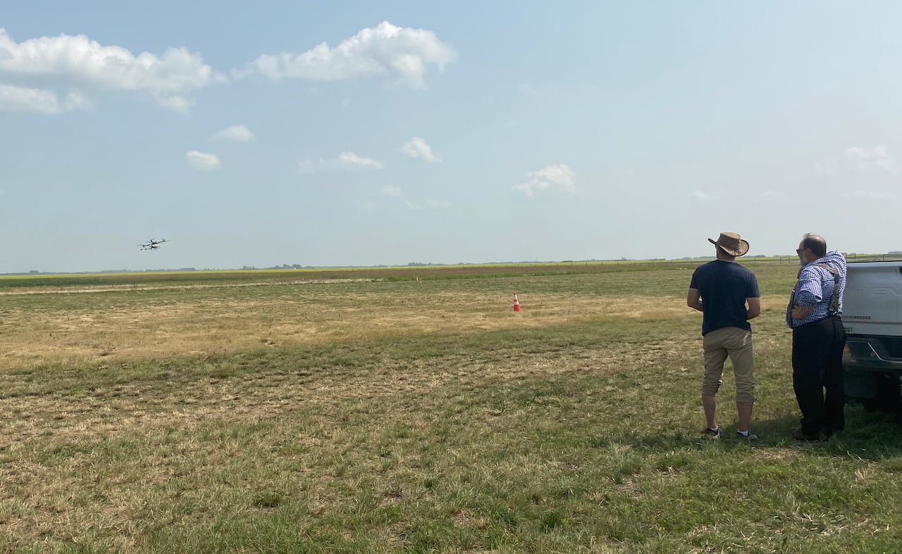 default-186 Two men stand near the back of a pickup truck in a field, watching a drone flying in the distance.